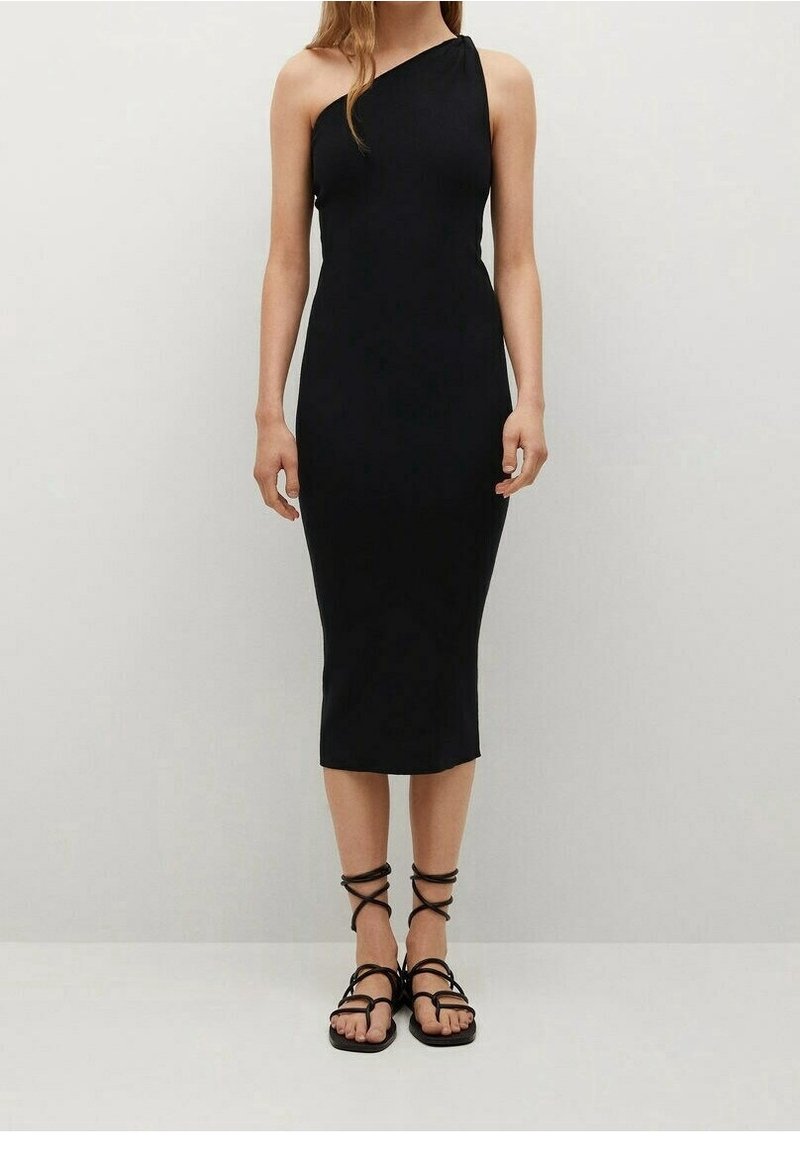 Femme portant une robe midi noire sans manches, à une épaule, et des sandales plates noires à brides, debout devant un fond clair uni.