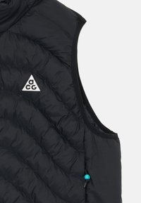 Melnā pūkainā veste ar augstu kaklu, kas rotāta ar kvadrātveida rakstu, triāngulāru logotipu un kontrastējošu zilu auklu detaļu apakšmalā.