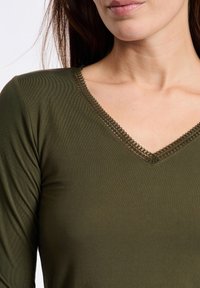 Maglietta verde oliva a maniche lunghe con scollo a V e dettagli in pizzo lungo il collo, realizzata in tessuto morbido e liscio.