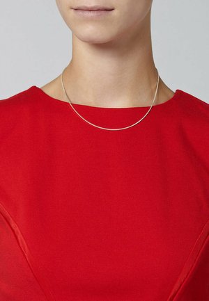 Eine silberne Halskette liegt auf einem roten Stoffoberteil. Sie besteht aus einer glatten, dünnen Kette mit einer zarten Textur und minimalistisches Design.