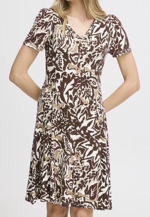 Femme portant une robe mi-longue à manches courtes avec un motif abstrait de feuilles et de fleurs marron et beige sur fond blanc.