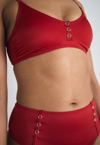 Crveni bikini set s visokim strukom i bralette topom. Značajne detalje čine tri srebrna prstena na sredini. Glatka tekstura.