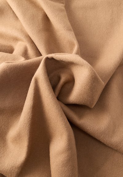 Pehmeä, sileä kamelinvärinen kangas, jossa näkyvät taitokset ja kiertyvä laskostus korostamassa rakennetta ja lempeitä varjoja.
