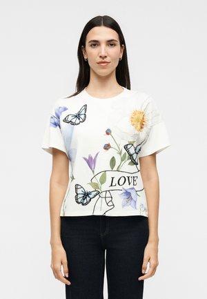 Kvinde med langt mørkt hår bærer en hvid t-shirt med farverige blomster, blå sommerfugle og ordet "LOVE" foran. Hun står med arme afslappet.