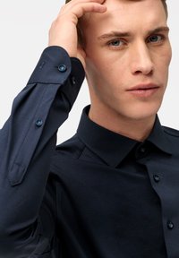 Chemise bleu marine avec col, manches longues et boutons bleus. Le tissu semble lisse avec un léger éclat. Le poignet a une piqûre d'accent.