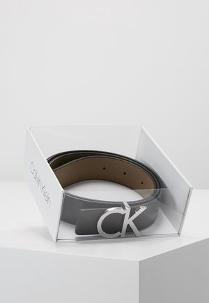 Zwinięty czarny skórzany pasek z srebrną klamrą "CK" w przezroczystym plastiku i białym pudełku z logo Calvin Klein na białej powierzchni.