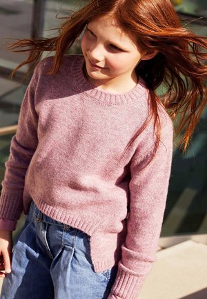 Una niña con un jersey de punto rosa y unos vaqueros azules está de pie al aire libre, con su largo pelo rojo ondeando al viento.