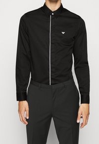 Camicia nera con bottoni, a righe verticali bianche contrastanti, maniche lunghe e un piccolo logo ricamato sul taschino sinistro.