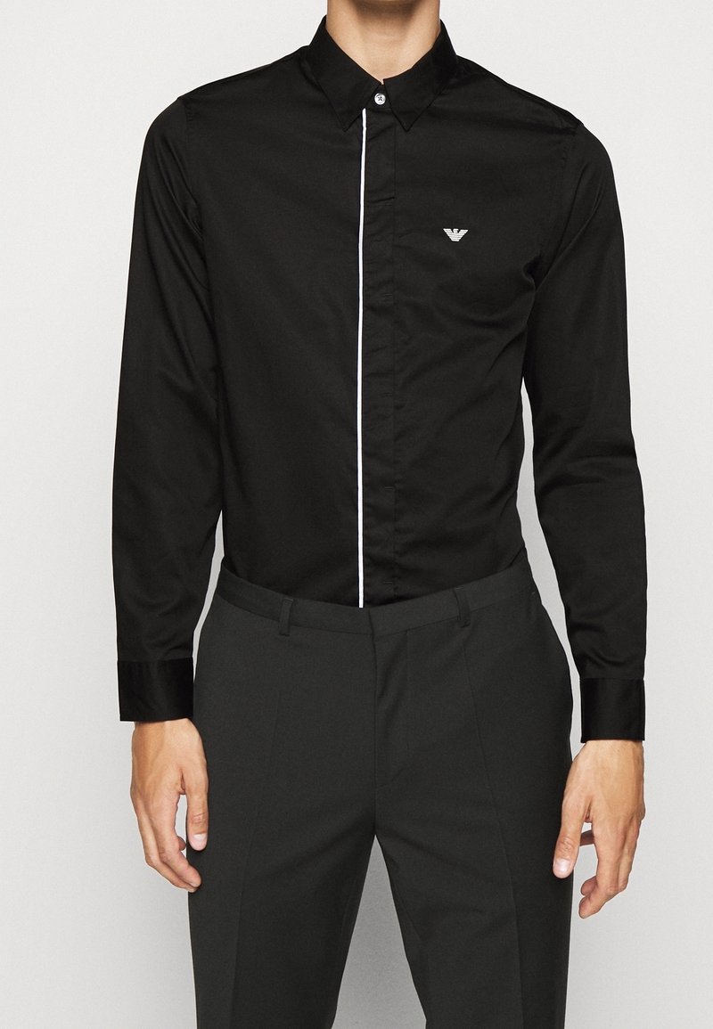 Camicia nera con bottoni, a righe verticali bianche contrastanti, maniche lunghe e un piccolo logo ricamato sul taschino sinistro.