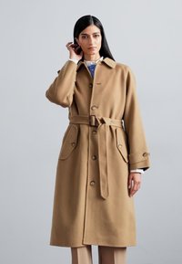 Beige wool trench coat with a belted waist, large side pockets, and a notch collar. Featuring button closure and long sleeves.