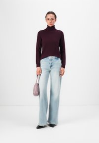 Burgundowy sweter polo, jasnoniebieskie szerokie dżinsy i czarne szpilki. Trzyma małą szarą torebkę. Gładka faktura, codzienny design.