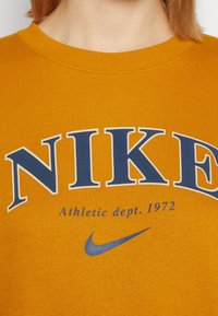 Sweatshirt Nike en jaune moutarde avec texte imprimé en bleu, comportant "Athletic dept. 1972" et un logo swoosh Nike. Tissu texturé.