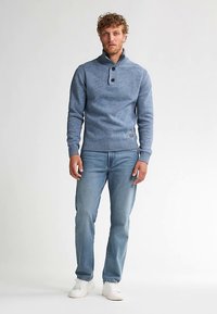 Hellblauer Strickpullover mit hohem Kragen, Drei-Knopf-Verschluss und gerippten Bündchen, kombiniert mit hellblauen Jeans und weißen Sneakern.