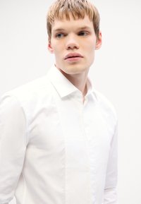 Chemise blanche avec un design à plis à l'avant, col pointu et fermeture à bouton. Tissu lisse avec un léger éclat et coupe ajustée.