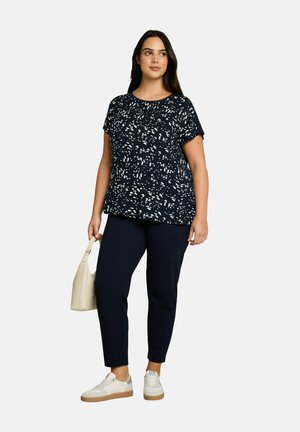 Kobieta ubrana w czarno-białą wzorzystą bluzkę z krótkim rękawem, czarne spodnie, białe tenisówki, trzyma jasnobeżową torebkę, stoi i patrzy w prawo.