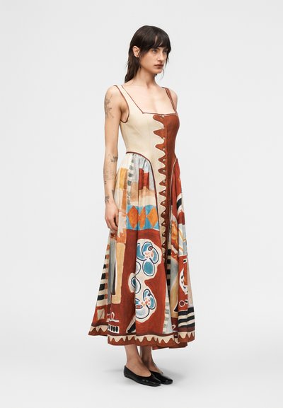 Γυναίκα που φοράει μίντι φόρεμα χωρίς μανίκια με τυπωμένα αφηρημένα μοτίβα σε καφέ, κρεμ, μπλε και πορτοκαλί, συνδυασμένο με μαύρες μπαλαρίνες.