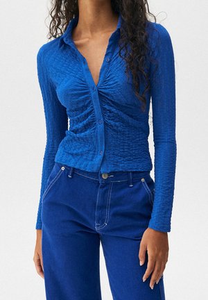 Frau trägt ein eng anliegendes, strukturiertes blaues Langarmhemd mit Knöpfen und hoch taillierte blaue Hose vor einem einfarbigen Hintergrund.