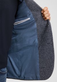 Graue texturierte Jackeninnenseite mit blauer Fütterung, ausgestattet mit einer kleinen Reißverschlusstasche und kontrastierenden Nähdetails. Hand teilweise sichtbar.