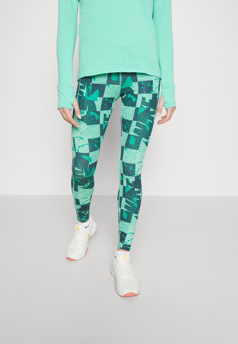 Haut de sport vert menthe à manches longues associé à des leggings teal à motifs avec des formes géométriques et des accents texturés. Baskets blanches.