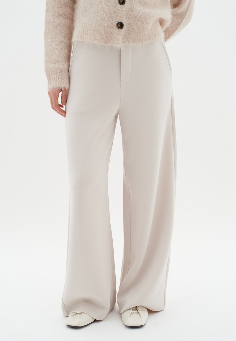 Pantalons à jambe large en tissu beige doux, avec une taille haute et des coutures minimalistes. Associés à un pull en doux rose clair et des chaussures blanches.