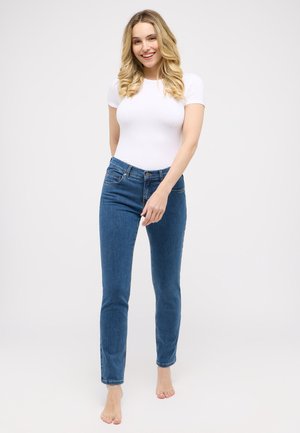 Schmale Jeans aus Denim in Blau, mit hoher Taille und einem Fünf-Taschen-Design. Kombiniert mit einem eng anliegenden weißen T-Shirt und barfuß.