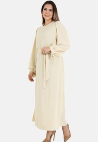 A long, cream-colored dress with puffed sleeves and a tied waist detail. The fabric has a soft texture, and the design features a straight cut.