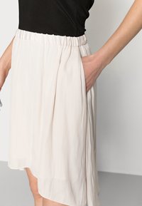 Femme portant une jupe beige clair avec une taille élastique et plaçant sa main dans la poche latérale, associée à un haut noir.