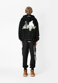 Persona de pie de espaldas a la cámara, vistiendo una sudadera con capucha negra con un gráfico de caricatura, pantalones negros con múltiples parches y botas naranjas.