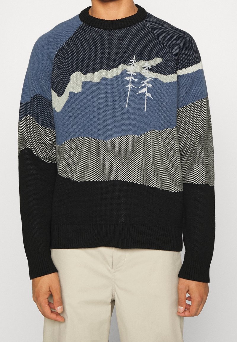 Pull en tricot pour hommes avec motifs de montagne bleus, gris et noirs et deux dessins d'arbres blancs sur la poitrine, assorti à un pantalon beige.