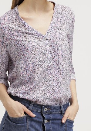 Vrouw draagt een lichtpaarse blouse met patroon en lange mouwen, ingestopt in een blauwe jeans met hoge taille, met handen in de zakken.