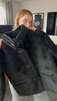 Jeune femme aux cheveux blonds tenant un manteau noir à boutons, duveteux, dans une pièce lumineuse avec des œuvres encadrées au mur.