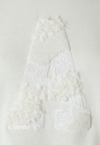 Bílá mikina s texturovaným písmenem "A" vyrobeným z chlupaté tkaniny a flitrů, se jemnými slonovinovými akcenty a hladkým pozadím.