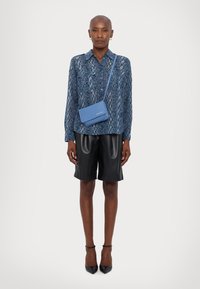 Blå skjorte med slangeskinnsmønster, svarte skinnshorts og en liten blå crossover-veske. Modellen har på seg svarte spisse hæler, noe som gir et elegant utseende.