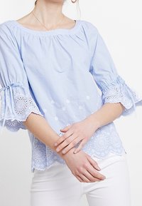 Light blue striped blouse with embroidered lace trim and bell sleeves. Features a rounded neckline and floral accents on the fabric.