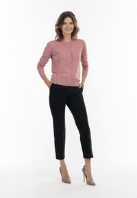 Cardigan button-up di un rosa chiaro, vestibilità rilassata, indossato sopra pantaloni scuri. Le caratteristiche includono polsini a coste e una superficie liscia. Tacchi alti neutri.