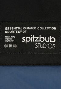 Schwarzer Stoffanhänger mit weißem Text, der "Essential Curated Collection" und "spitzbub STUDIOS" liest. Hat eine strukturierte Oberfläche und Nähdetails.