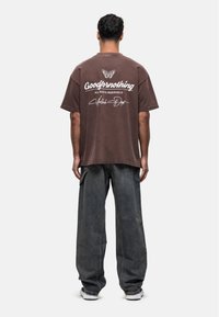 Bruine oversized T-shirt met witte tekst en vlinderafbeelding op de achterkant, gecombineerd met grijze baggy jeans en witte sneakers.