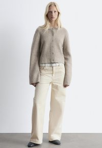 Brauner, taillierter Strickpullover mit Knopfverschluss, kombiniert mit hochgeschnittenen, hellen, weit geschnittenen Cordhosen und dunklem Schuhwerk.