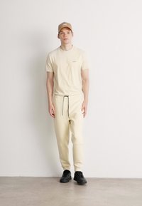 Beige T-Shirt und passende Jogginghose mit Kordelzug, kombiniert mit einer sandfarbenen Mütze. Das Modell trägt schwarze Sneakers und steht vor einem neutralen Hintergrund.