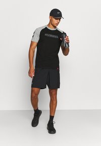 Svart og grå atletisk t-skjorte med korte ermer, logo på forsiden, kombinert med svarte shorts og svarte joggesko. Holder en grå sportsflaske.