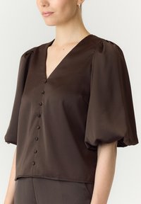 Femme portant un chemisier marron foncé avec des manches bouffantes et un décolleté en V, avec une rangée de boutons recouverts sur le devant.