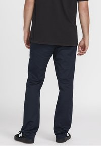 Pantalon bleu marine avec une coupe droite, présentant une texture lisse et une taille élastique. Porté avec des chaussures noires affichant des accents blancs.