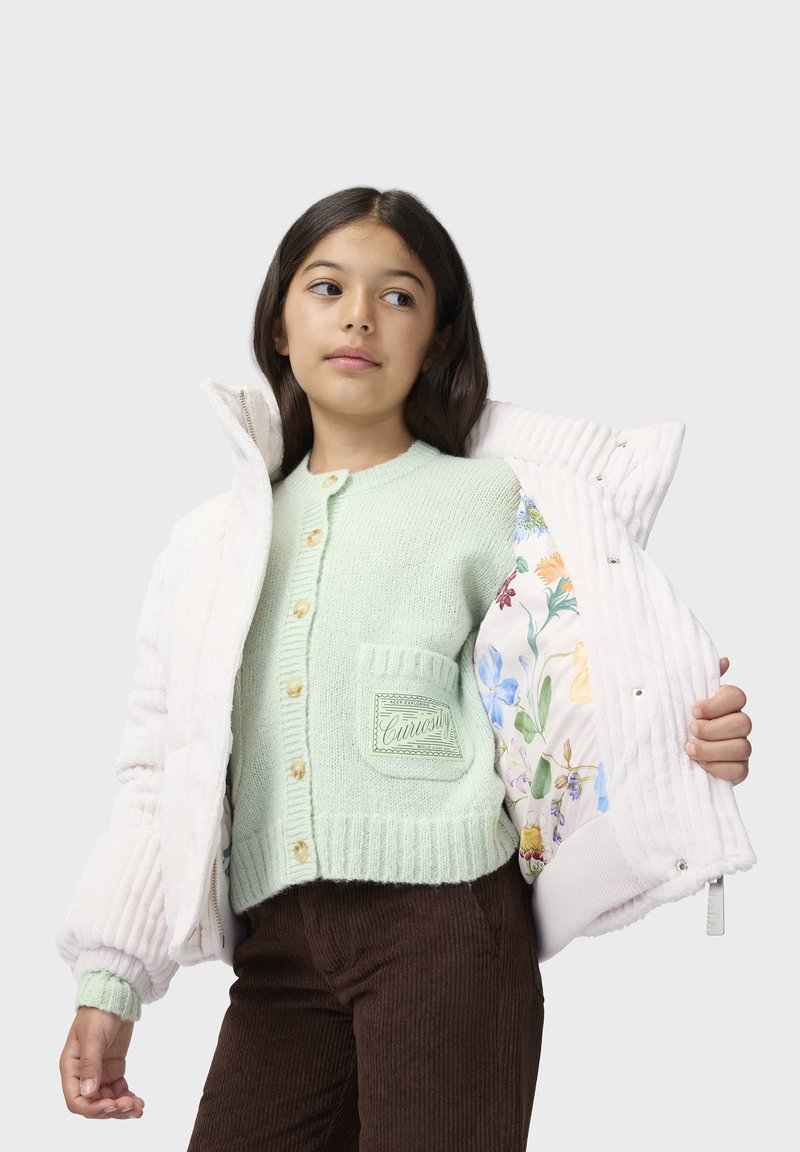 Fille en pantalon en velours côtelé marron et cardigan vert menthe montrant une veste matelassée blanche avec une doublure florale colorée.