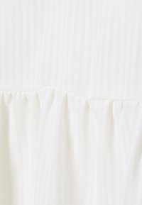 Tessuto bianco texturizzato con righe verticali e un dettaglio di ruffle arricciato nella parte inferiore. Materiale morbido e leggero con una leggera lucentezza.