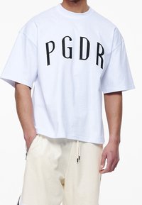 Mężczyzna w białej, oversize'owej koszulce z czarnym napisem "PGDR" oraz kremowych szortach na sznurki z zasuwakami.