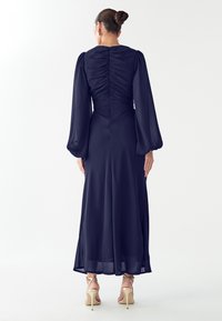 Tmavě modré dlouhé šaty s řaseným zadním dílem, nafouklými rukávy a splývavou sukní. Mají průsvitnou látku a jsou kombinovány s béžovými sandály na podpatku.
