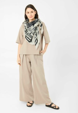 Frau trägt beige weite Hosen und passendes lockeres Oberteil, schwarz-weiß gemusterten Schal und schwarze Riemchensandalen, steht mit der Hand in der Tasche.