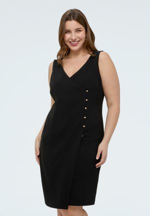 Smilende kvinde iført en ærmeløs sort slå-om-kjole med guldknapper ned langs den ene side, stående mod en ensartet lys baggrund.