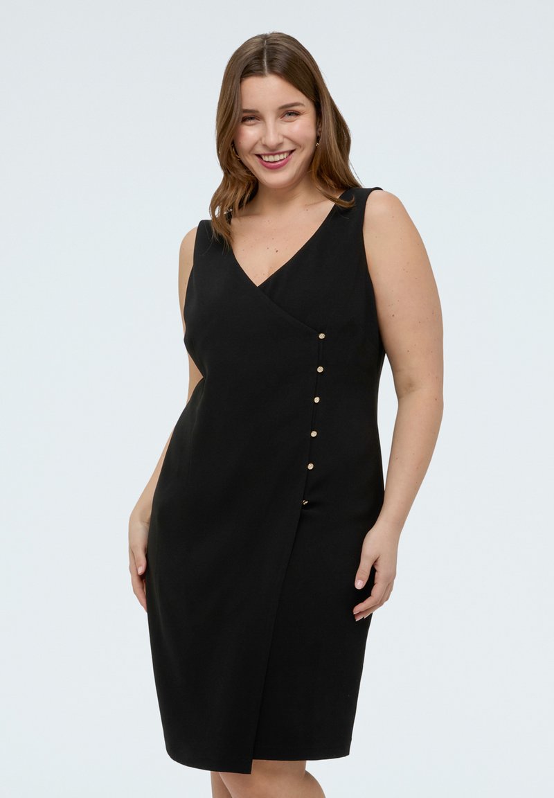 Hymyilevä nainen, joka pukeutunut hihattomaan mustaan kietaisumekkoon, jossa on kultaiset napit toisella sivulla, seisoo yksivärisen vaalean taustan edessä.