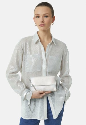Sheer silver shirt with two front pockets and a collared neckline. Holding a metallic silver clutch with a chain strap. Blue jeans.